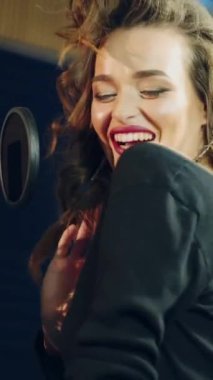 Harika bir kadın kayıt stüdyosunun arka planında profesyonel bir mikrofonun önünde neşeyle dans edip şarkı söylüyor. Mutlu müzisyen mikrofonun başında dikey müzik videosu yapıyor.