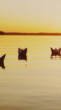 Akşam güneşinin sarı ışınları gün batımında nehirde yüzen kağıt teknelerin üzerinde parlıyor. Origami gemileri açık havada dikey görüntülere yansıyor.