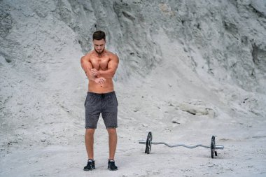 Kaslı adam plajda halterle egzersiz yapıyor. Sağlıklı yaşam tarzı ve spor kavramı.