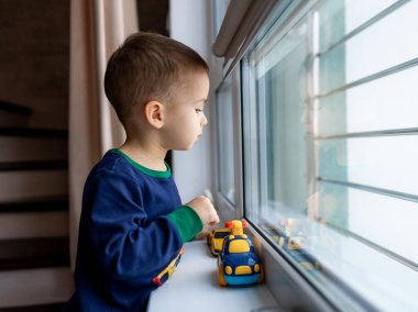 Küçük çocuk pencerenin yanında duruyor ve elinde oyuncaklarla pencereden dışarı bakıyor.