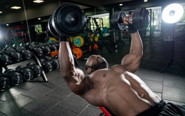 Spor salonunda dambıllarla egzersiz yapan kaslı bir adam.