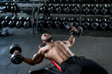Spor salonundaki halterlerle egzersiz yapan kaslı, üstsüz Afrikalı Amerikalı adam.