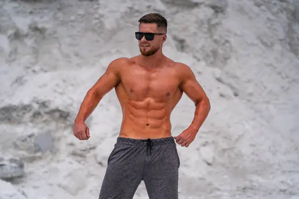 Handsome muscular man with stylish haircut and beard posing on the beach showing his abs and torso