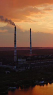 Gün batımında nehrin yanındaki fabrikada. Gece gökyüzüne karşı tüten piposu olan endüstriyel fabrika. Ekolojik sorunlar. Hareket kamerası geri. Hava görüntüsü. Dikey video