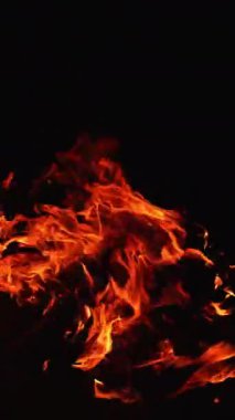 Büyük şenlik ateşinde yanan odun parçaları. Uzun alev havaya doğru uzanıyor sola doğru eğiliyor. Arkaplanda karanlık var. Dikey video