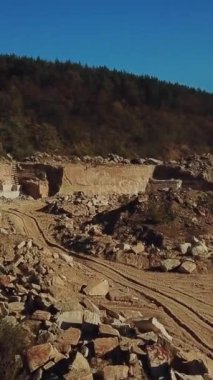 İnsanlar, yüksek ve uzun bir taş ocağının yakınında, üzerinde taş kayalar ve mavi gökyüzünün arka planında sıcak bir yaz günü üzerinde kozalaklı bir orman bulunan dev taş yığınları etrafında çalışıyorlar. Hava görüntüsü. Kamera ileri doğru hareket ediyor. Dikey video