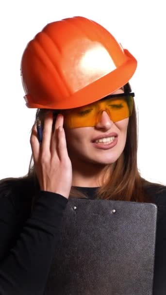 オレンジ色のヘルメットと保護眼鏡の美しい女性は 彼女の手に黒いプランチとペンを保持し 白い背景に何かについて話します バーティカルビデオ — ストック動画