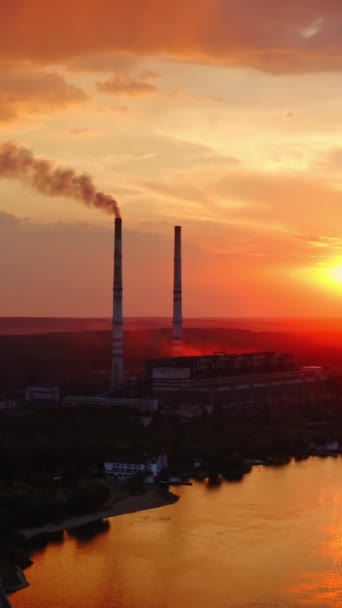 オレンジの日没の産業企業 川の近くにパイプが付いている大きい植物 煙突から空中に汚れた煙が吸い込まれる 環境汚染 空からの眺め バーティカルビデオ — ストック動画