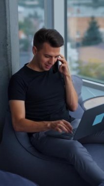 Genç bir erkek, aynı anda bilgisayar ve telefon kullanıyor. Pencere kenarında rahat bir yerde çalışan serbest çalışan meşgul bir adam. Yüksek açı. Dikey video