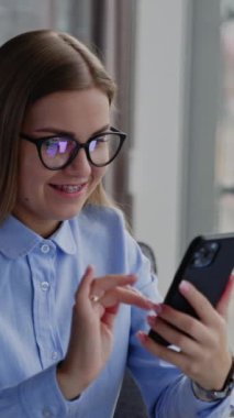 Ofisteki genç bayan masada oturuyor. Gözlüklü kadın dizüstü bilgisayarın önünde ve telefonuna mesaj yazıyor. Kapatın. Dikey video