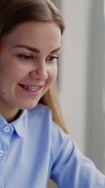 Ofisteki masada diş teli takmış neşeli, güzel bir genç bayan. Dişi, dizüstü bilgisayara bakarak not alıyor. Ofis içi arka plan. Dikey video