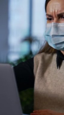 Koruyucu maskeli kadın çalışan dizüstü bilgisayarında rahatsız görünüyor. Kadın bilgisayarı kapatır, maskesini çıkarır ve kameraya gülümser. Dikey video
