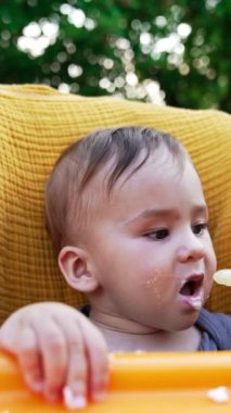 Güzel, aç, beyaz bir bebek, yulaf lapalı bir kaşığa kendi rızasıyla ağzını açar. Annesinin küçük oğlunu dışarıda beslemesi. Bulanık doğa arkaplanı. Dikey video