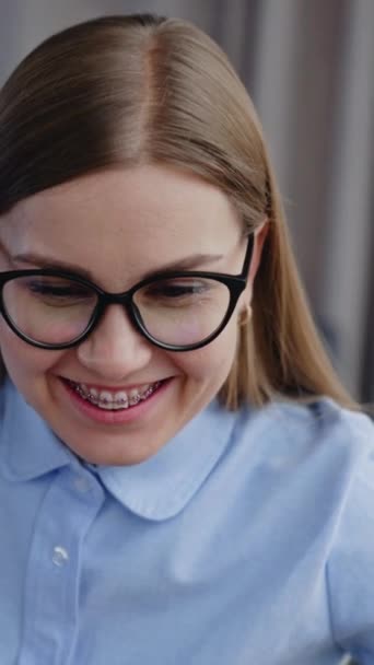 快乐地微笑着 长发女士戴着眼镜 牙齿上戴着牙套 看着电脑屏幕 办公室里快乐的员工 垂直录像 — 图库视频影像