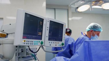 Ameliyat sırasında hastanın yaşam parametrelerini gösteren monitörler. Doktorun ekibi arka planda çalışıyor..