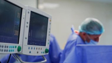 Ameliyat altındaki hastanın yaşam belirtilerini gösteren iki ekran. Tıbbi ekip arka planda bulanık çalışıyor.