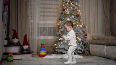 Sevimli çocuk elinde bir emzik tutarak odanın önünden geçiyor. Çocuk kanepede duruyor. Arkaplanda Noel ağacı.