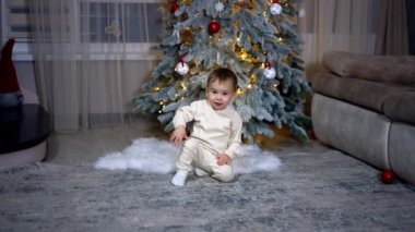 Odada Noel ağacının yanında oturan küçük sevimli melek bebek. Sevimli çocuk sanki bir şey bekliyormuş gibi hareketsiz oturuyor ve kamera yaklaşırken sevimli bir şekilde gülümsüyor..