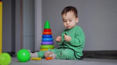 Sevimli çocuk elinde ayakkabı bağcığıyla yerde oturuyor ve ağzına alıyor. Oyuncaklar bebeğin etrafında. Kapalı arkaplan.