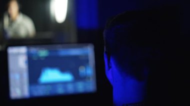 Profesyonel stüdyoda bilgisayar ekranının önünde çalışan bir ses mühendisinin dikiz görüntüsü. Düet arka planda bulanık bir pencerede icra eder.