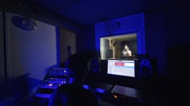 Modern profesyonel müzik stüdyosu. Ses mühendisi ekipmanın önünde oturuyor. Erkek şarkıcı yan odada, pencerenin arkasında bir şarkı söylüyor..