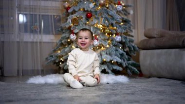 İnanılmaz sevimli küçük çocuk mutlu bir şekilde gülüyor ve kameraya doğru sürünüyor. Arkaplanda süslenmiş Noel ağacı.