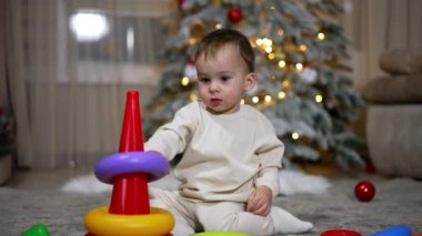 Güzel, sakin bir erkek bebek yerde oturan oyuncak bir piramitle oynuyor. Arka planda Noel için dekore edilmiş oda.