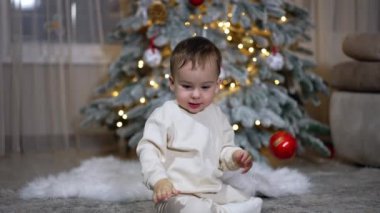 Beyaz elbiseli küçük sevimli çocuk yerde oturuyor. Bebek kırmızı topu onun önüne attı. Arkaplanda Noel ağacı.