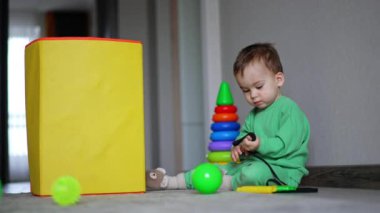 Huzurlu erkek bebek yerde oturuyor. Çocuk kordonu elinde geriyor. Oyuncak kutusu ve oyuncaklar etrafa saçılmış..