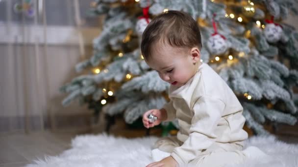 可爱的小男孩快乐地玩玩具 身穿白色西服的孩子坐在白色柔软蓬松的格子布上 一边玩着圣诞树上的玩具 — 图库视频影像