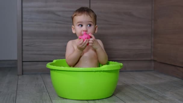 ウォッシュボウルに座っているゴムピンクのおもちゃで遊ぶ美しい赤ちゃんの少年 子供は水からおもちゃを取り出し それを彼の口に引き込む — ストック動画