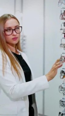 Optik dükkanındaki genç sarışın. Zarif elbiseli çekici bir kadın gözlükleriyle bükülüyor. Hareket eden kamera var. Dikey video