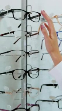 Gözlük, optik mağazadaki standın üzerinde. Modaya uygun gözlükleri seçen genç alıcı kadın. Göz giyimi konsepti. Dikey video