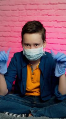 Çocuk izole edilmiş, koruyucu steril tıbbi maskeyle evde oturuyor. Yüzünde ve eldivenlerinde. Karantina, enfeksiyonu önleyin. Koronavirüsü durdurun. Evde kal Dikey video