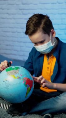 Kendini izole etmiş bir çocuk, Covid-19 'u öğrenen küreye bakıyor. Tıbbî maskeli bir çocuk. Gezegeni düşünüyor. Karantina, karantina ve sosyal uzaklık konsepti.