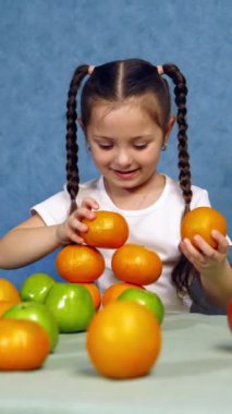 Taze meyvesi olan tatlı küçük bir kız. Masaya portakal kulesi yapan komik kız. Çocuk mavi arka planda organik meyvelerle oynuyor. Dikey video