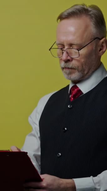 一个严肃的白发男人的画像 有一个文件夹 戴眼镜的老人穿着西装在文件夹上写字 老人在黄色背景下表现出不同的面部情绪 垂直录像 — 图库视频影像