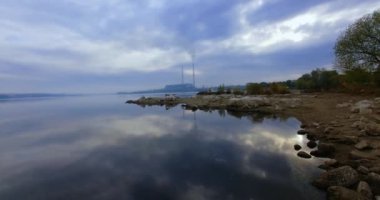 Nehrin taşlı kıyısında bitkiler yetişmiyor. Gri bulutlar suyun pürüzsüz yüzeyine yansıyor. Arkaplanda sanayi fabrikası boruları.