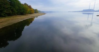 Bulutlu gri bir günde, hava aracı nehrin ayna yüzeyinde uçuyor. Ağaçların büyüdüğü kumlu bankanın manzarası. Arka planda sisli ufuk.