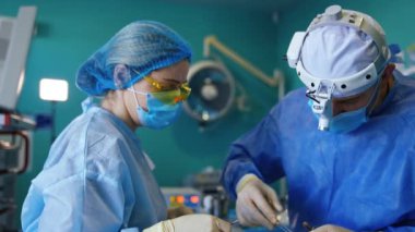 Beyaz erkek cerrah hastayı dikkatlice dikiyor. Operasyon prosedürünün son aşaması. Hemşire metal aletleri doktora veriyor..