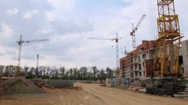 Çok katlı binanın şantiyesine birkaç vinç yerleştirildi. Zeminde çeşitli yapı malzemeleri var. Arka planda büyüyen ağaçlar.