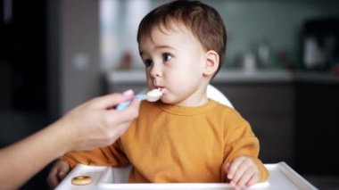 Güzel beyaz erkek bebek evde yemek yiyor. Çocuk annesinden bir kaşık alıyor ve ağzı doluyken dikkatlice kameraya bakıyor..
