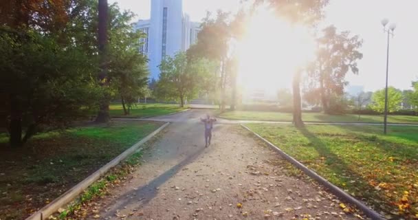 太陽の光の中で公園の道を歩いている小さな少年 カメラは子供から距離を置いて 彼は追いつくために走り始めます — ストック動画