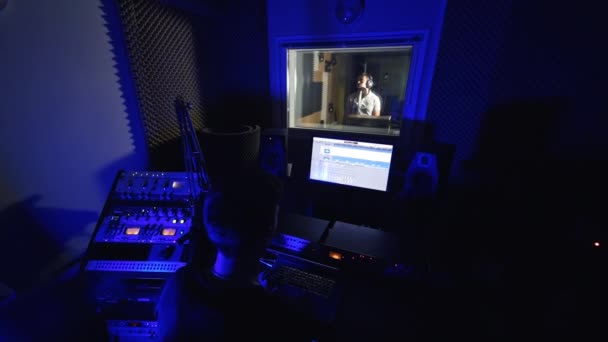 小さな暗いレコーディングルームで働く音楽プロデューサー サウンドエンジニアはコンピュータの画面を見ています バックドロップで歌う男性歌手 — ストック動画