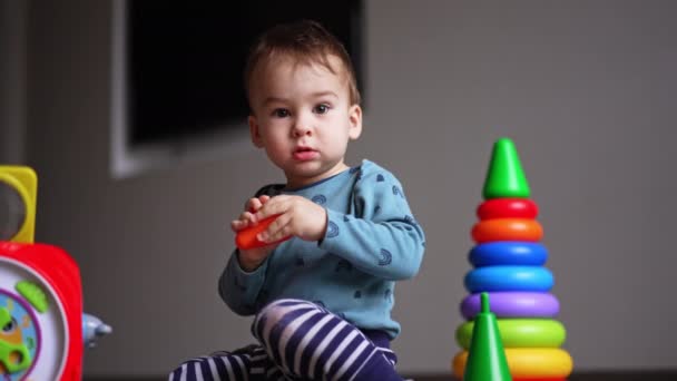 おもちゃの間で床に座っている赤ん坊の少年がカメラを見つめている トッドラーはプラスチックのおもちゃで遊んで手で絞っている ローアングルビュー — ストック動画