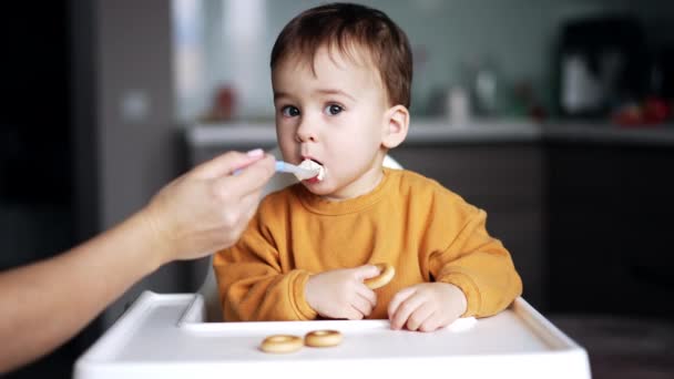 オレンジ色のシャツの小さな男の子は バッグで遊ぶテーブルに座っています 母親は息子の口にスプーンを閉じ 子供は袋に焦点を当てています — ストック動画