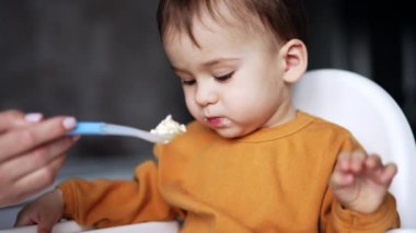 Küçük oğlan beslenme koltuğunda oturuyor. Annem kaşığı oğluna yulaf lapası verir, o da alır ve elini öne uzatır..