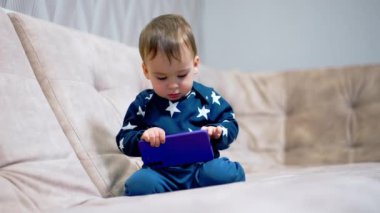 Oturma odasındaki kanepede oturan sevimli çocuk. Mavi elbiseli sevimli çocuk telefonuyla oynuyor ve dikkatlice inceliyor..