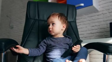 Siyah deri sandalyede oturan küçük, ciddi bir çocuk. Güzel çocuk uzaktan kumandayı elinde tutuyor ve düğmelere basıyor.