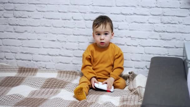 可爱的孩子靠着白色的砖墙坐在床上 小孩拿着玩具手机看着摄像机 — 图库视频影像